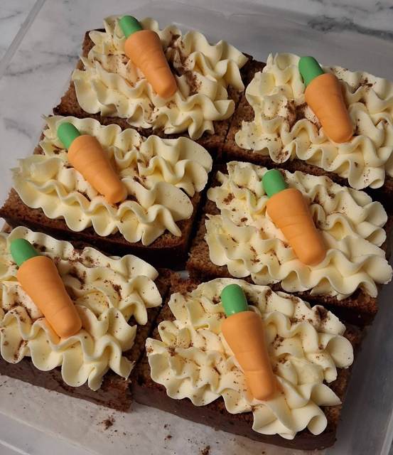 Six squares of carrot cake topped with buttercream and marzipan carrots